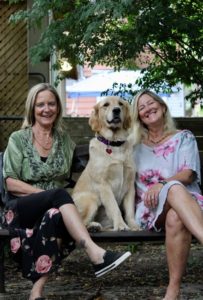 Trish Drynan, golden retriever Suki, and Andrea Drynan all smiling and sitting on a bench